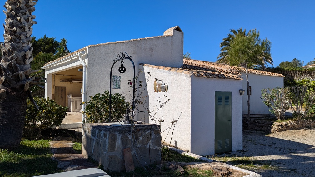 Exterior view of a traditional Mediterranean outbuilding featuring white facades, terracotta roof tiles, and a decorative stone well with wrought iron details.