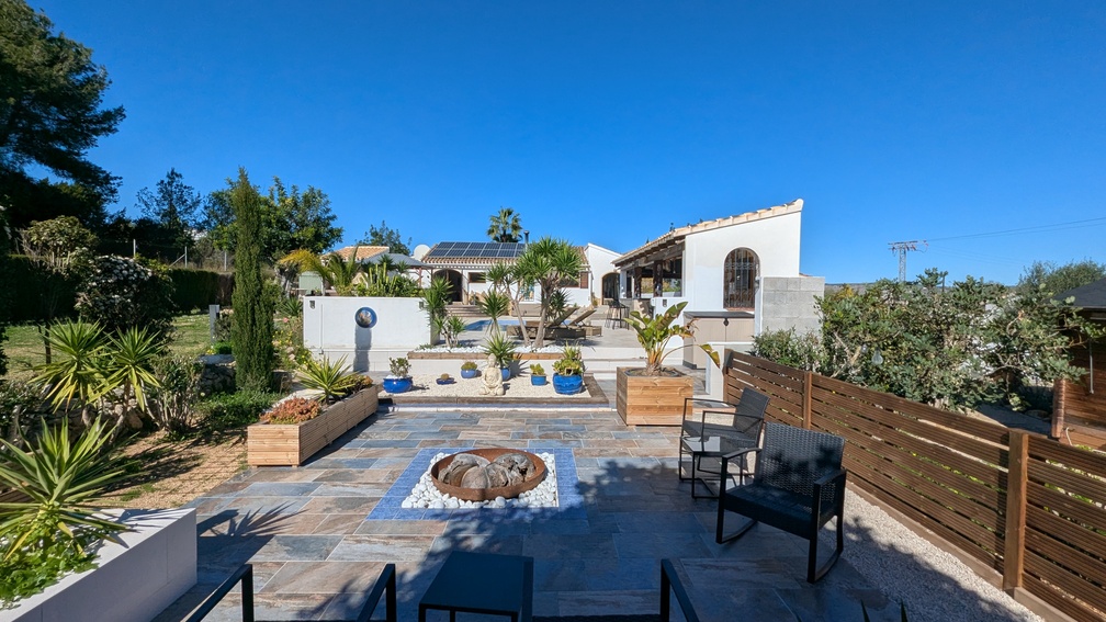 Exterior view of the landscaped terrace featuring a central fire pit, slate paving, and steps leading to the main villa with rooftop solar panels.