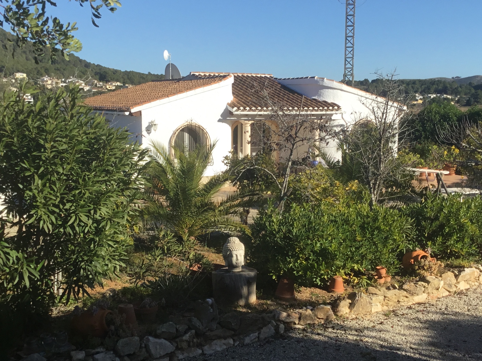 Exterior view of the white-washed Mediterranean villa featuring arched windows, terracotta roof tiles, and a landscaped garden with palm trees and a Buddha statue.