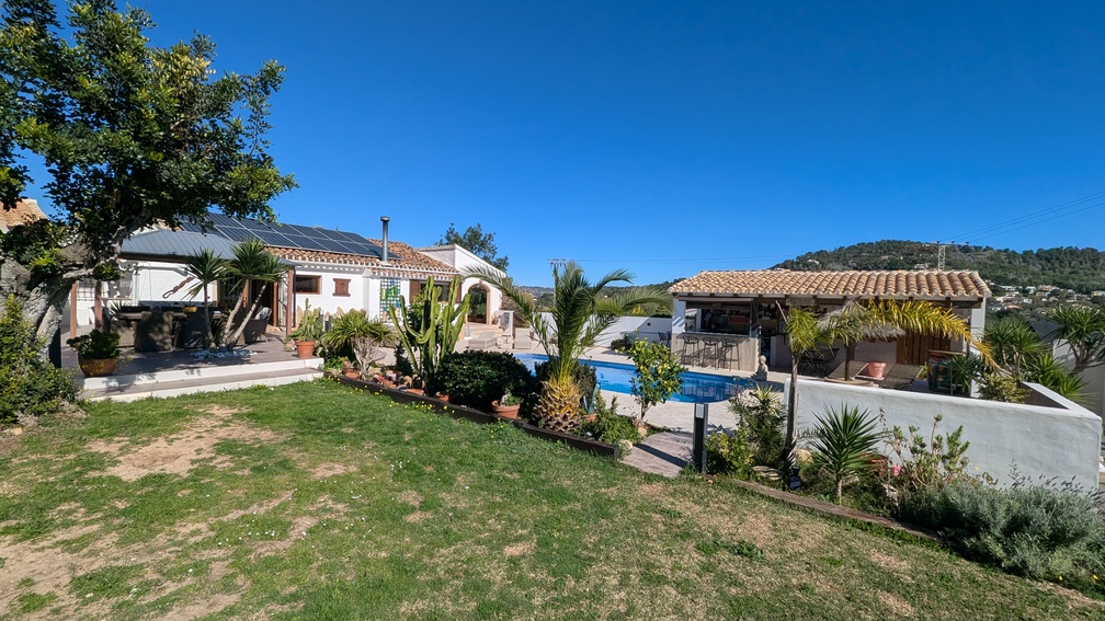 Exterior view showing the main house with solar panels, a private pool, and a covered summer kitchen/bar area surrounded by Mediterranean gardens.