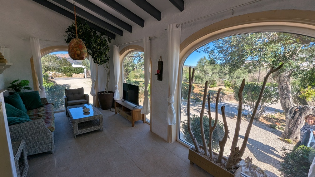 Glazed naya featuring traditional dark wooden beams, arched windows with countryside views, and a comfortable seating area with wicker furniture and tiled flooring.