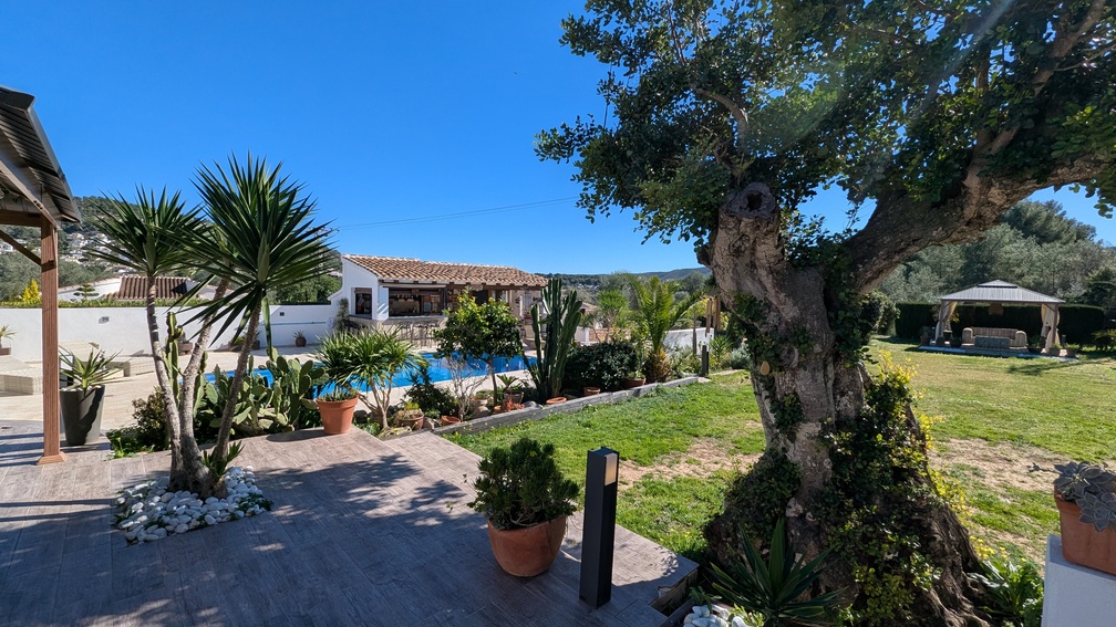 Landscaped Mediterranean garden featuring a private pool, a fully equipped summer kitchen with a tiled roof, and a shaded gazebo on a manicured lawn.