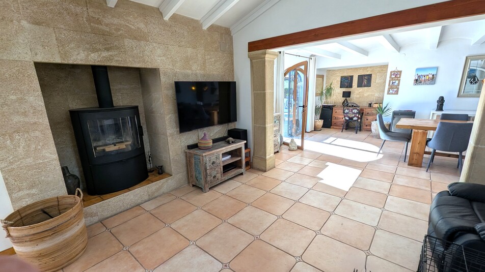 Living area featuring a modern wood-burning stove recessed into a natural stone wall, terracotta tile flooring, and exposed white ceiling beams.