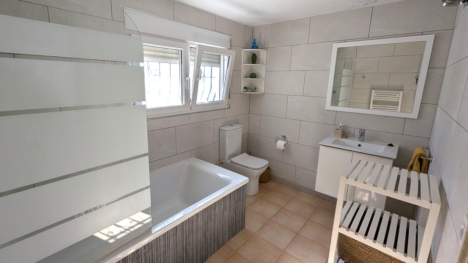 Modern bathroom featuring a built-in bathtub with a frosted glass screen, a white vanity unit, and neutral large-format wall tiles.