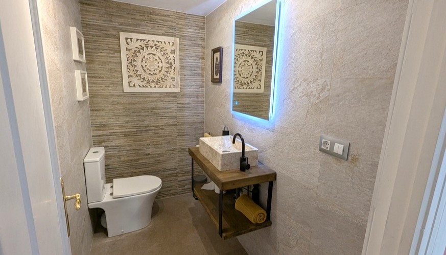Modern guest bathroom featuring a stone vessel sink on a floating wooden vanity, a backlit LED mirror, and textured stone-effect wall tiling.