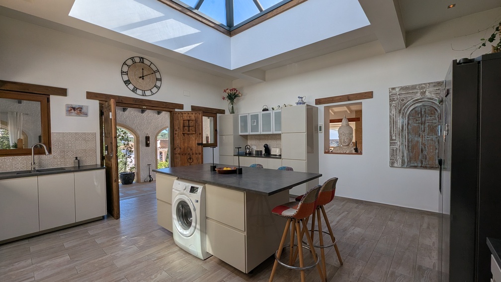 Modern kitchen featuring a central island and a large glass skylight. Traditional wooden doors and lintels lead to the arched naya, blending style with heritage.