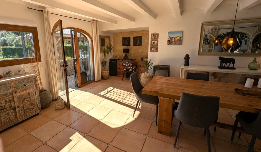 Open-plan dining area featuring exposed white beams, terracotta flooring, and a solid wood table. An arched glass door provides direct access to the garden terrace.
