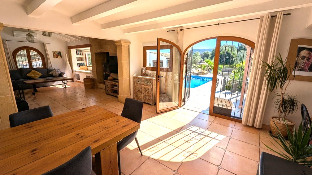 Open-plan living and dining area featuring white ceiling beams, Tosca stone pillars, and arched glass doors opening directly to the pool terrace.