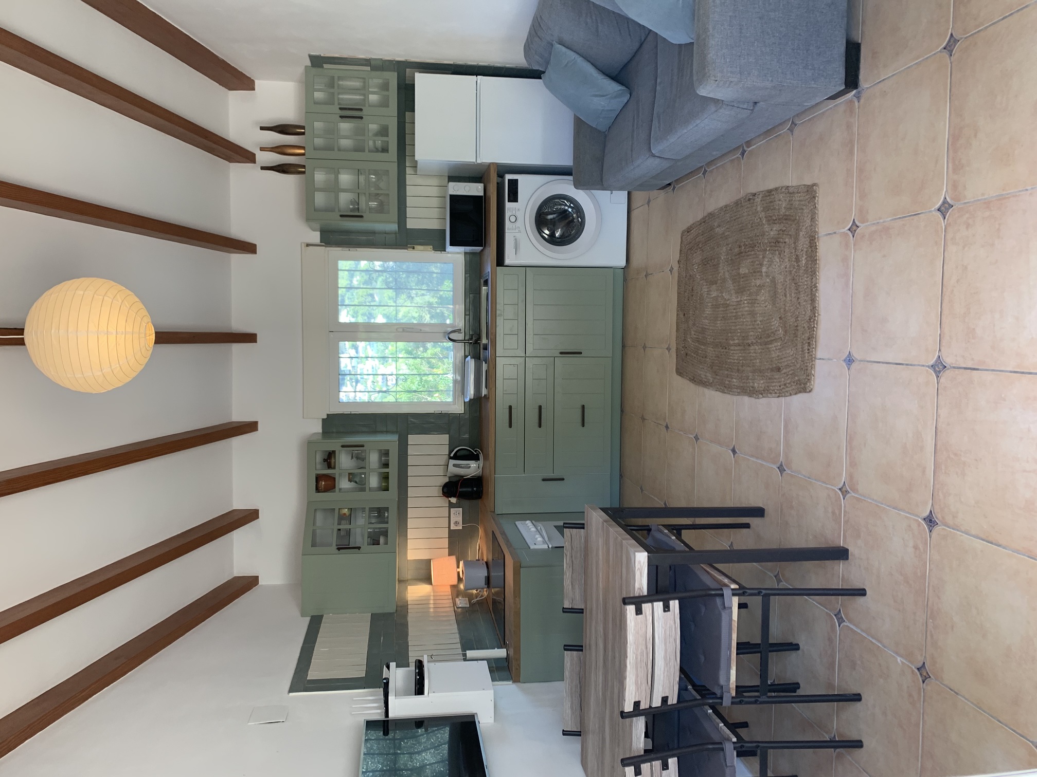 Open-plan living space featuring a sage green kitchen with wooden worktops, exposed ceiling beams, and a dining area set on traditional terracotta floor tiles.