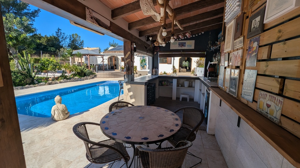 Outdoor bar and summer kitchen with exposed wooden beams, mosaic dining table, and direct views of the private pool and landscaped garden.