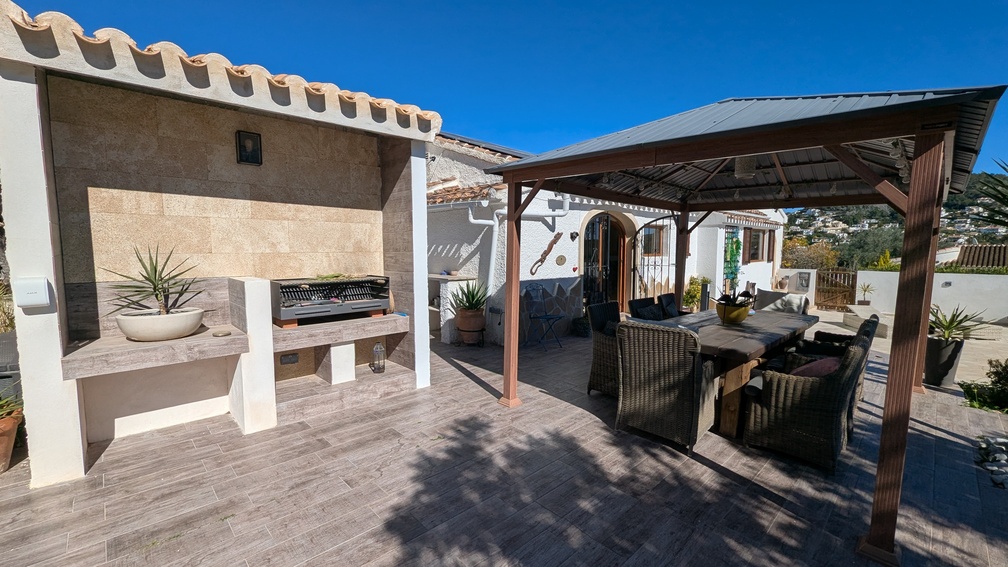 Outdoor dining area featuring a built-in stone BBQ station and a large gazebo-covered terrace with wicker seating on wood-effect tiled flooring.