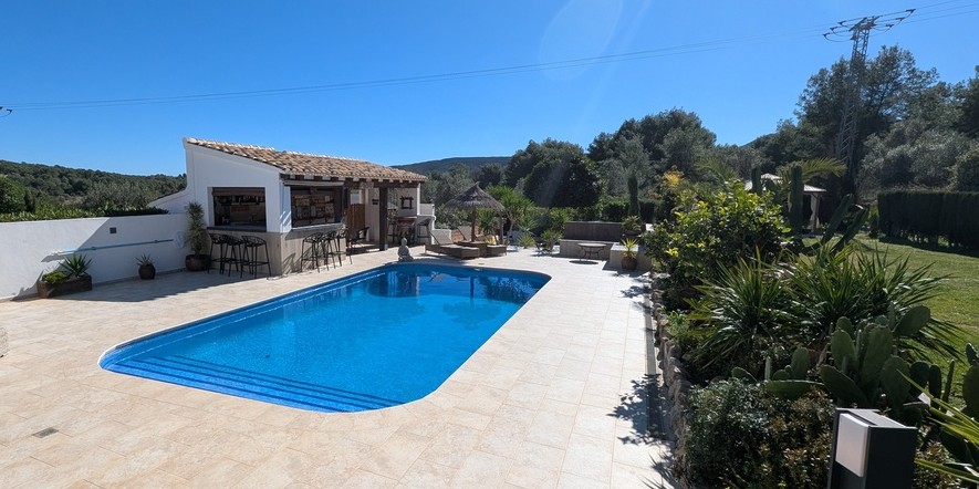Outdoor swimming pool area featuring a stone-tiled terrace, a fully equipped summer kitchen with a bar, and landscaped gardens with Mediterranean plants.