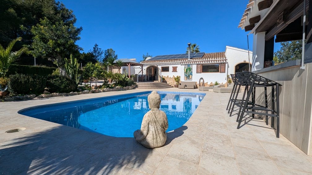 Private pool area with stone tiling, featuring a Buddha statue, a modern outdoor bar with stools, and a villa equipped with solar panels.