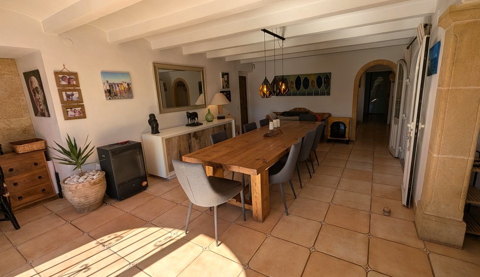 Spacious dining area featuring a large solid wood table, terracotta tile flooring, and a white beamed ceiling. A Tosca stone pillar and arched doorway highlight the traditional finca character.