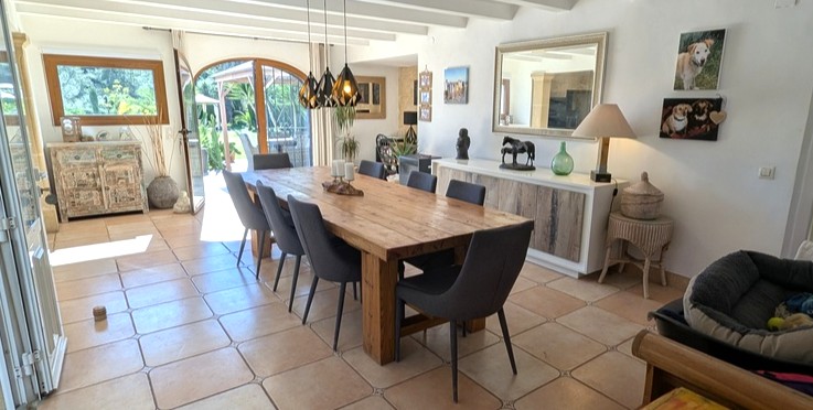 Spacious dining area featuring a rustic wooden table, contemporary seating, and terracotta flooring under a beamed ceiling, with direct access to the garden through arched doors.