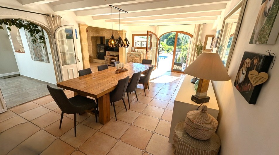 Spacious dining area featuring a solid wood table, exposed white ceiling beams, and terracotta flooring. Arched glass doors provide direct access to the pool terrace.