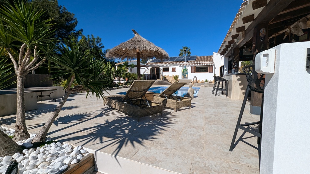 Sun-drenched terrace with stone tiling, featuring wicker loungers, a tiki-style parasol, and a private pool adjacent to the villa equipped with rooftop solar panels.