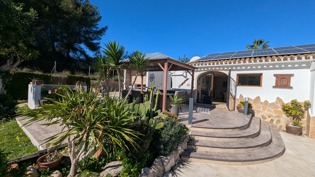 Terrace with curved stone steps, a wooden gazebo for outdoor dining, and a white facade featuring solar panels and Mediterranean landscaping.