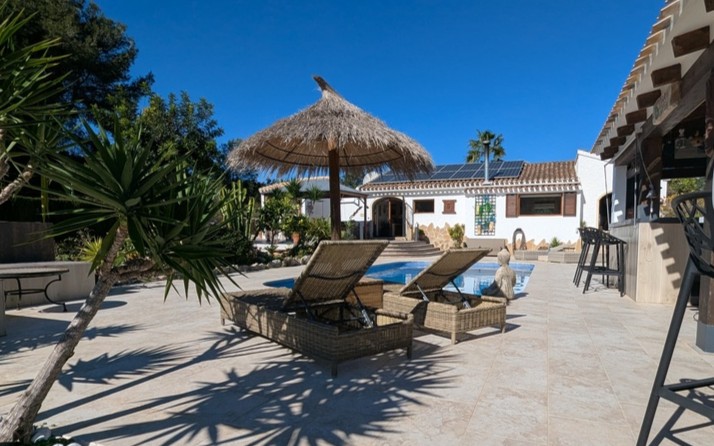 Tiled terrace featuring a private pool, wicker loungers under a thatched umbrella, and a summer bar. The main house includes roof-integrated solar panels.