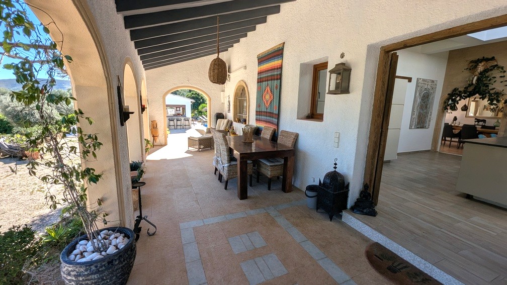 Traditional Mediterranean naya featuring dark wooden beams, arched openings, and a dining area. This shaded terrace connects the interior to the garden and summer kitchen.