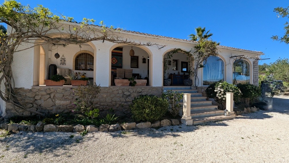 Traditional Mediterranean villa facade featuring an arched naya porch, natural stone base, and climbing vines against a white exterior.