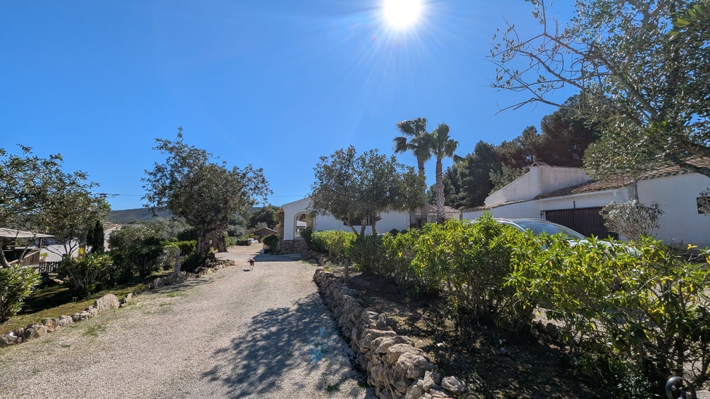 Wide gravel driveway lined with traditional stone walls leading to the white finca-style main house, surrounded by Mediterranean trees and palm trees.