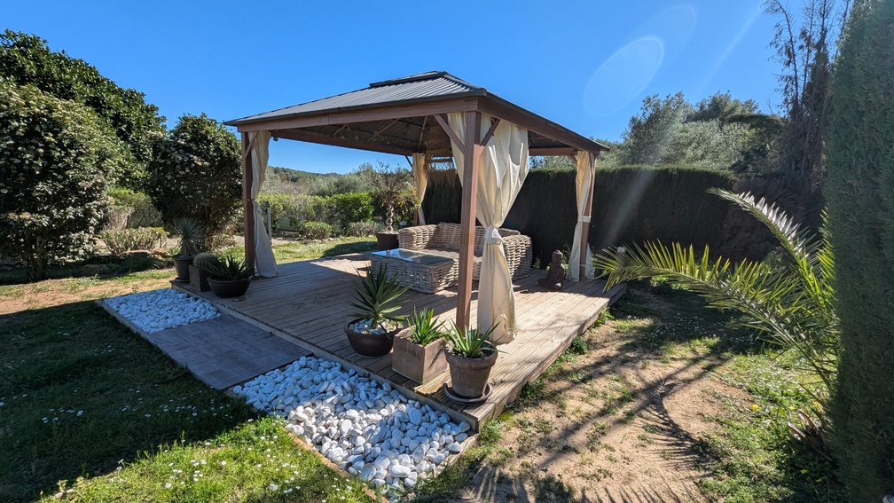 Wooden garden gazebo on a timber deck featuring wicker lounge furniture, cream curtains, and decorative white pebbles, set within a landscaped Mediterranean garden.