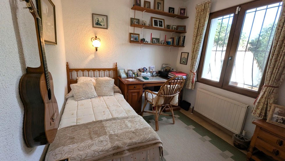 Bedroom featuring a single wooden bed, a classic desk with a wicker chair, and wall-mounted shelving. It includes a radiator and a window with security bars.