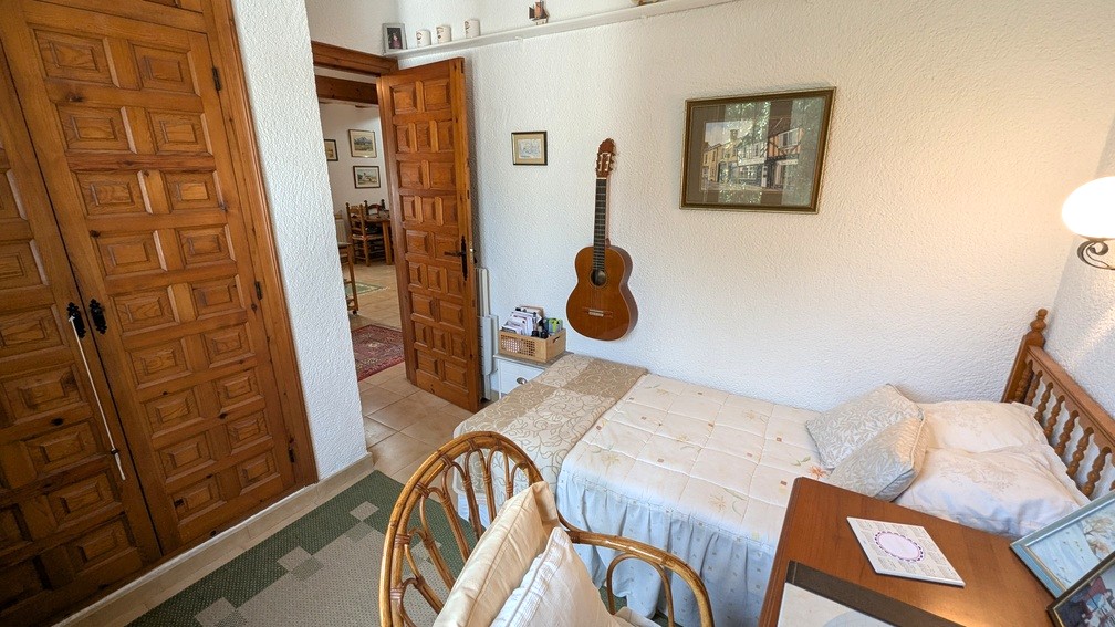 Bedroom with a single bed, built-in wooden wardrobes with traditional paneling, and white textured walls. An open door reveals the hallway and living space beyond.