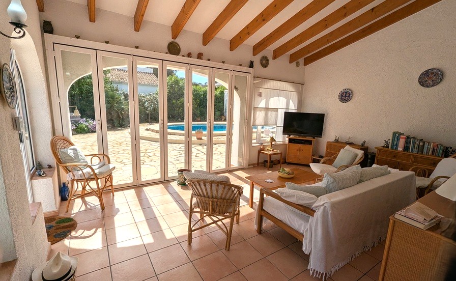 Bright living room featuring exposed wooden ceiling beams, terracotta flooring, and large folding glass doors opening onto the pool terrace.