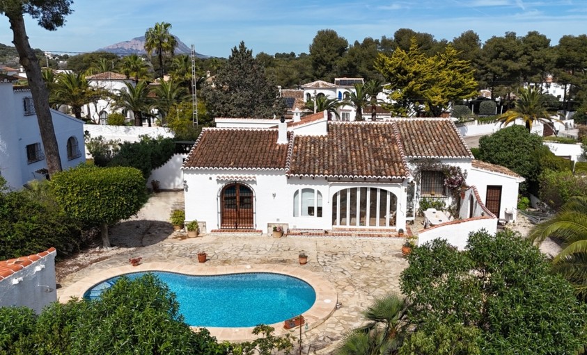 Elevated view of the villa featuring a kidney-shaped pool, stone-paved terrace, and traditional terracotta roof with the Montgó mountain in the distance.