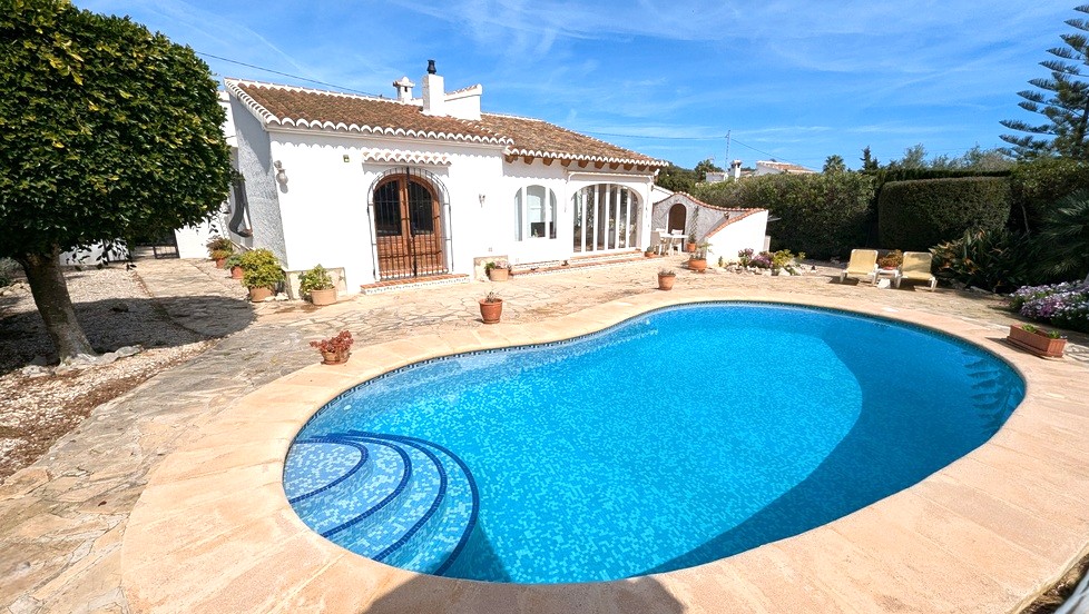 Exterior view of the kidney-shaped pool with blue mosaic tiling, surrounded by a stone terrace and the traditional white villa with terracotta roof.