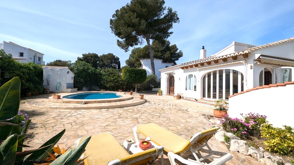 Exterior view of the Mediterranean villa featuring a kidney-shaped pool, natural stone terrace, and white facade with traditional wooden beams and arched glass doors.