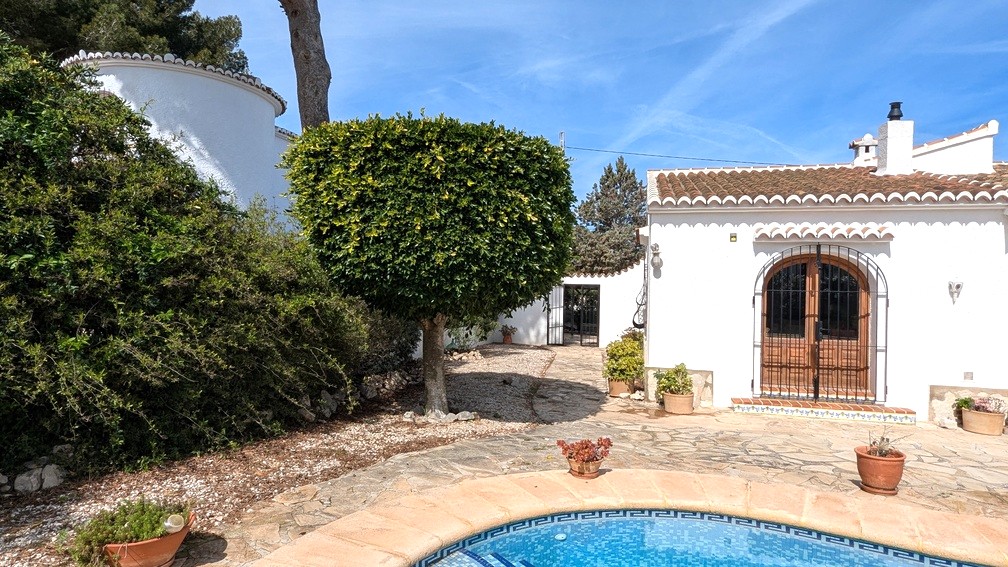 Exterior view of the villa featuring a swimming pool with mosaic tiling, a flagstone terrace, and traditional white-washed walls with arched wooden doors.