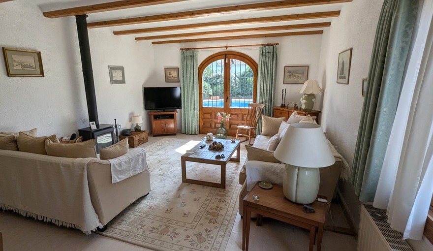 Living room with exposed wooden beams, a wood-burning stove, and arched wooden doors opening to the pool area. Traditional furniture and neutral tones define the space.