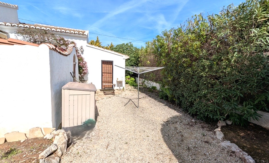 Side utility area with gravel flooring, featuring a rotary clothes dryer, storage shed, and privacy hedging next to the white-washed villa walls.