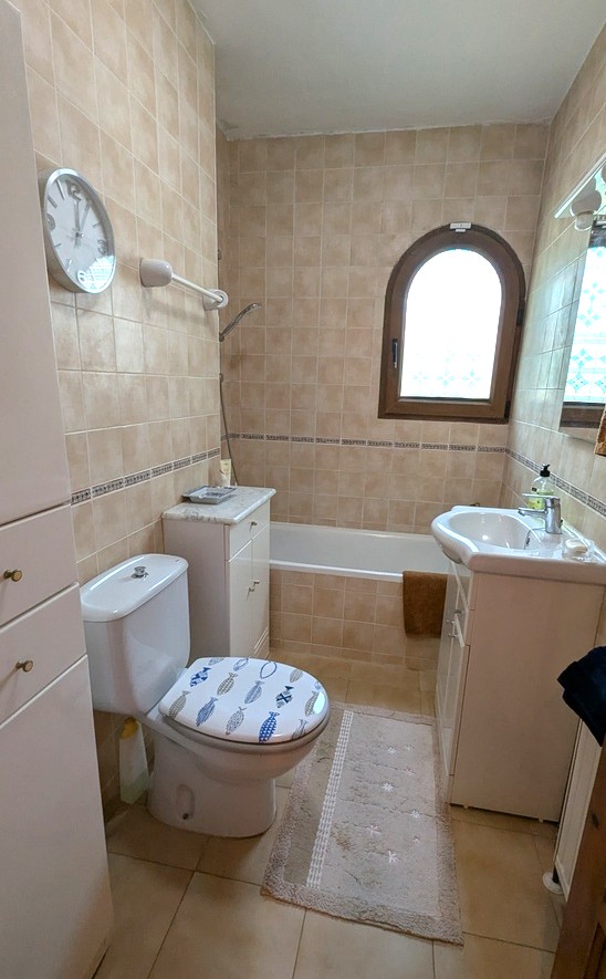 Traditional bathroom featuring beige tiling, a built-in bathtub, and a classic arched wooden window. Includes a white vanity, storage cabinets, and a decorative toilet lid.