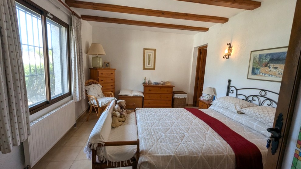 Traditional bedroom featuring exposed wooden ceiling beams, a wrought iron bed frame, and classic wooden furniture. Natural light enters through the window.