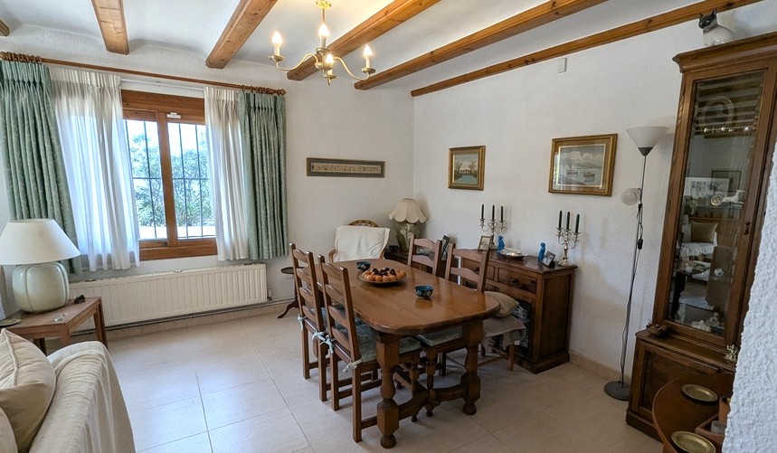 Traditional dining area featuring exposed wooden ceiling beams, a solid wood table set, and a classic sideboard. Natural light enters through a window with security bars.