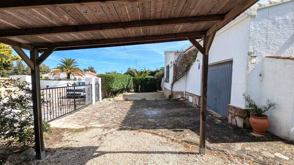 View from the wooden carport showing the stone-paved driveway, garage entrance, and wrought iron gate leading to the street.