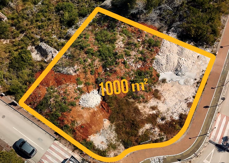 Aerial view of the 1,000 m² corner plot in Jávea, featuring cleared terrain with reddish soil and direct access to paved roads and urban infrastructure.