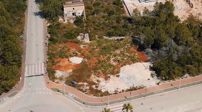 Aerial view of the 1,000 m² corner plot in Villas del Vent, showing the cleared terrain with red soil, surrounding pine trees, and paved infrastructure.