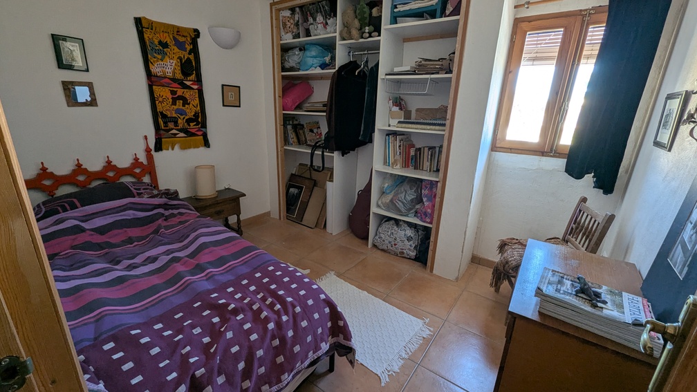 Bedroom featuring terracotta floor tiles, built-in open shelving, and a wooden window frame. The space includes a single bed and a wooden desk under natural light.