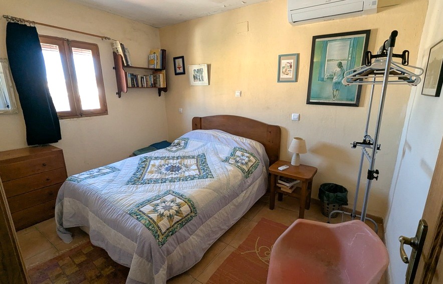 Bedroom with wooden furniture, tiled floors, and wall-mounted air conditioning. Features a classic patterned quilt, wall shelving, and natural light from a wooden-framed window.