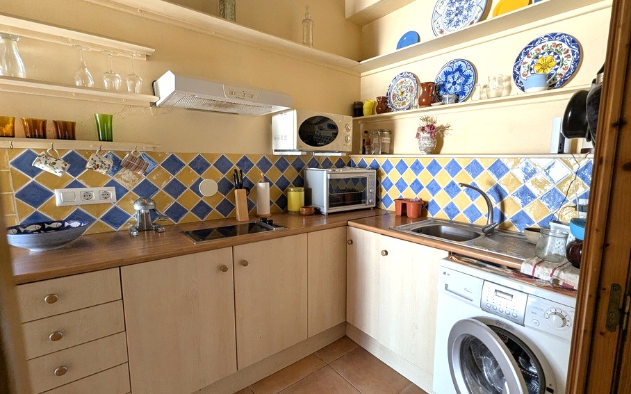 Compact kitchen featuring a blue and yellow diamond tile backsplash, wooden countertops, and open shelving with traditional ceramics. Includes a washing machine and appliances.
