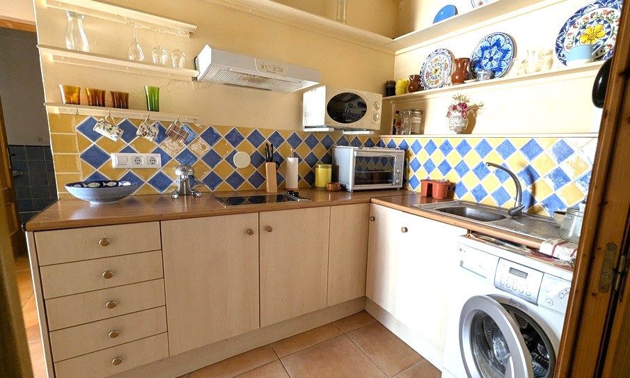 Compact L-shaped kitchen featuring a blue and yellow diamond tile backsplash, wooden countertops, and open shelving displaying traditional ceramics.
