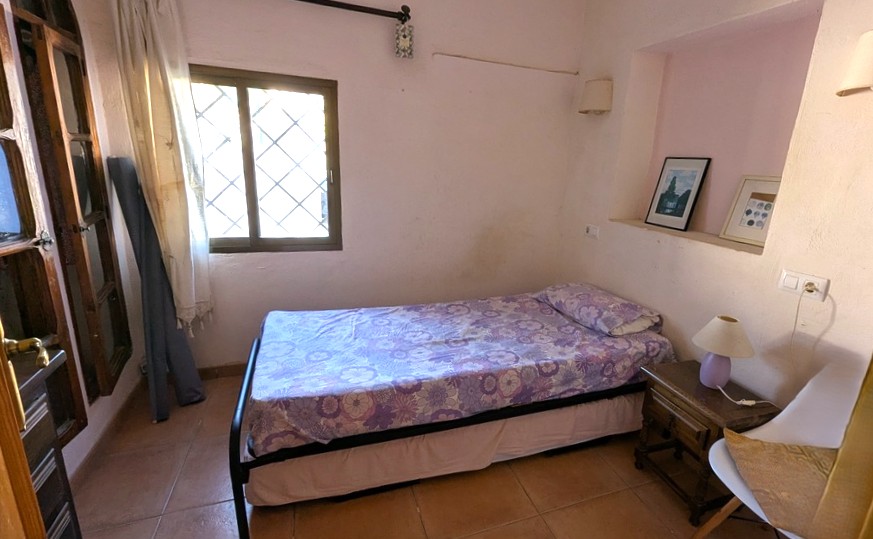Cozy bedroom featuring a single bed, terracotta tile flooring, and a recessed wall niche for decor. A window with traditional security bars provides natural light.