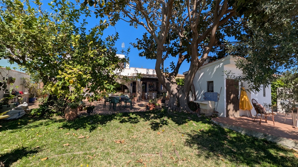 Mediterranean garden featuring mature trees, a lemon tree, and a lawn. Includes a paved patio with dining furniture and a white outbuilding near the traditional villa facade.