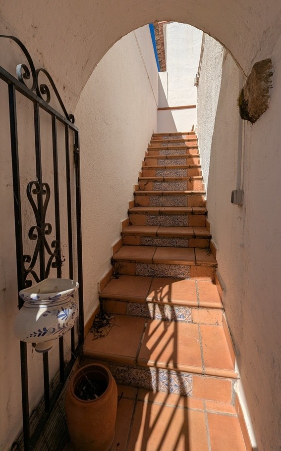 Narrow outdoor staircase featuring terracotta tiles and decorative ceramic risers, accessed through a white arched entrance with a wrought iron gate.