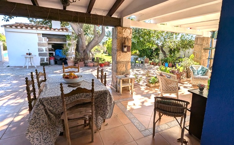 South-facing naya featuring Tosca stone pillars, exposed wooden beams, and a tiled dining area overlooking the garden and outdoor summer kitchen.